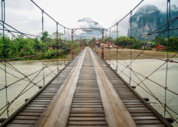 Wooden suspension bridge over river with misty mountains in Vang Vieng, Laos.
