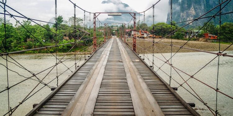 Wooden suspension bridge over river with misty mountains in Vang Vieng, Laos.