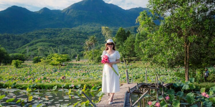 Woman in a white dress holds pink lotus flowers in a scenic Hoi An field, under a clear sky.