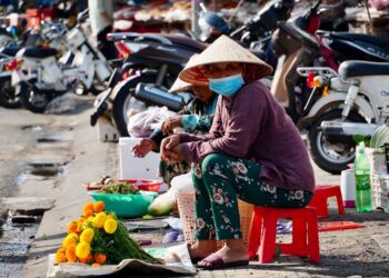Asian market vendor wearing traditional hat and mask sells vibrant marigold flowers on a sunny street.