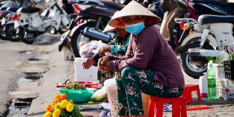 Asian market vendor wearing traditional hat and mask sells vibrant marigold flowers on a sunny street.