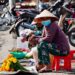 Asian market vendor wearing traditional hat and mask sells vibrant marigold flowers on a sunny street.