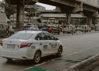 White taxi on a busy street in Quezon City, Philippines, with cars and overpasses in the background.