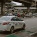 White taxi on a busy street in Quezon City, Philippines, with cars and overpasses in the background.