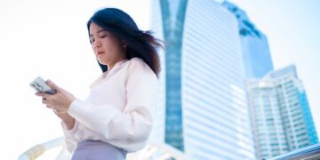 Asian woman using smartphone in Bangkok with modern skyscrapers in the background.