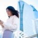 Asian woman using smartphone in Bangkok with modern skyscrapers in the background.