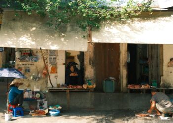 Two vendors at a busy Vietnamese street market selling goods under a canvas shade.